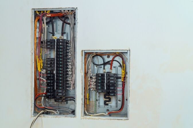 Two open electrical breaker panels with visible wiring and circuit breakers mounted on a white wall.