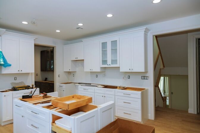 A kitchen under renovation with white cabinets being installed, tools and open drawers visible, and unfinished walls and surfaces.