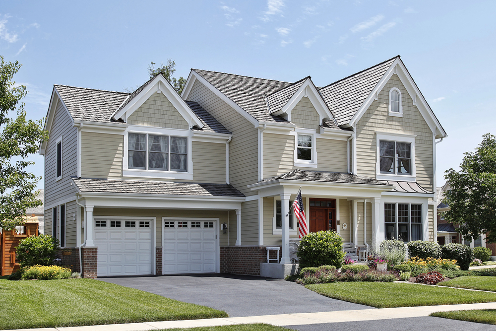 Two-story suburban house with beige siding, white trim, and a double garage. An American flag is displayed near the front porch, surrounded by landscaped gardens and a paved driveway.