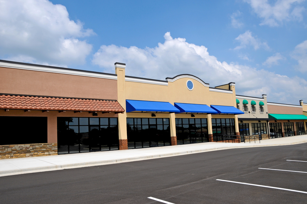 A row of empty retail storefronts with colorful awnings in a modern strip mall, viewed from an empty parking lot under a partly cloudy sky.