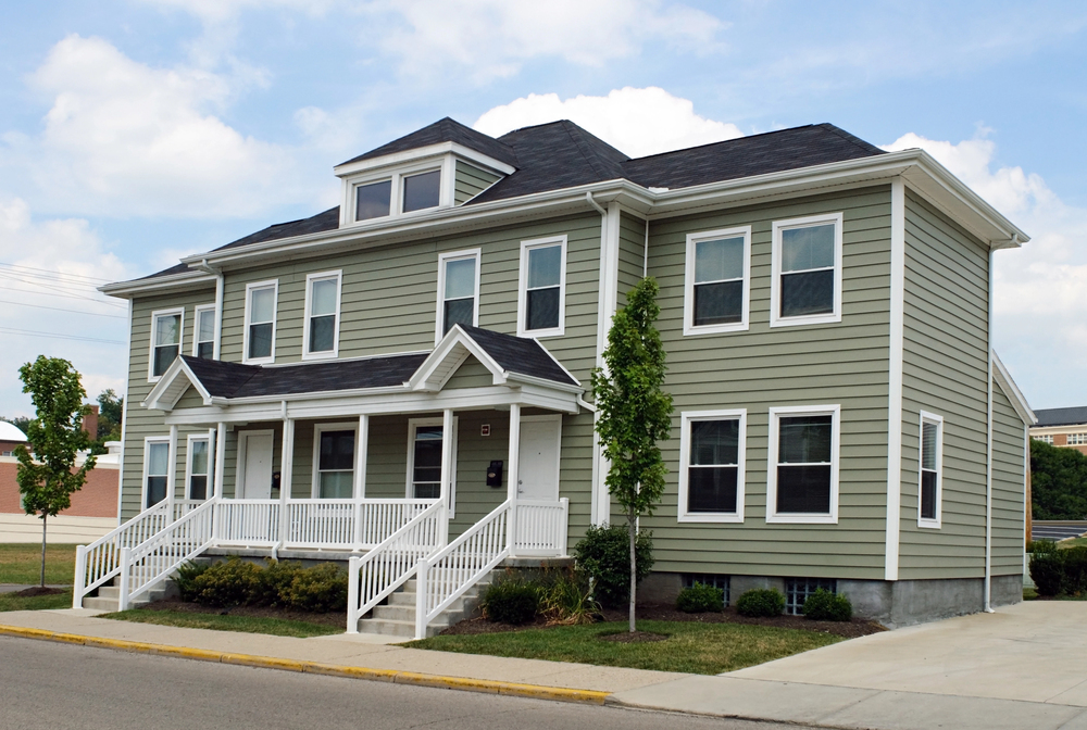 A two-story suburban duplex with gray siding, white trim, and two front porches, situated on a corner lot with small trees and shrubs.