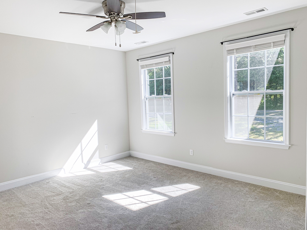 Empty room with light gray walls, beige carpet, two large windows with white blinds, and a ceiling fan; sunlight streams in through the windows.