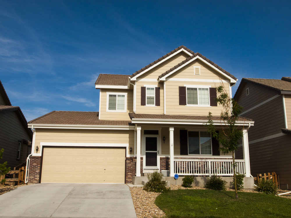 Two-story beige suburban house with a front porch, attached two-car garage, and a small front yard under a clear blue sky.