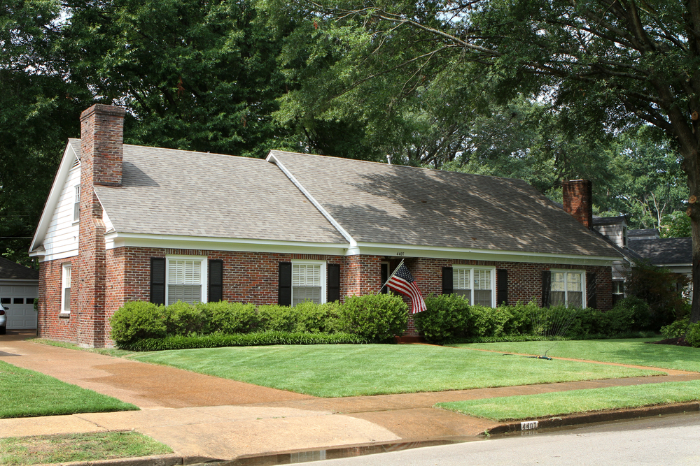Single-story brick house with gray roof, white trim, black shutters, and a front yard with neatly trimmed grass and bushes; an American flag hangs near the entrance.